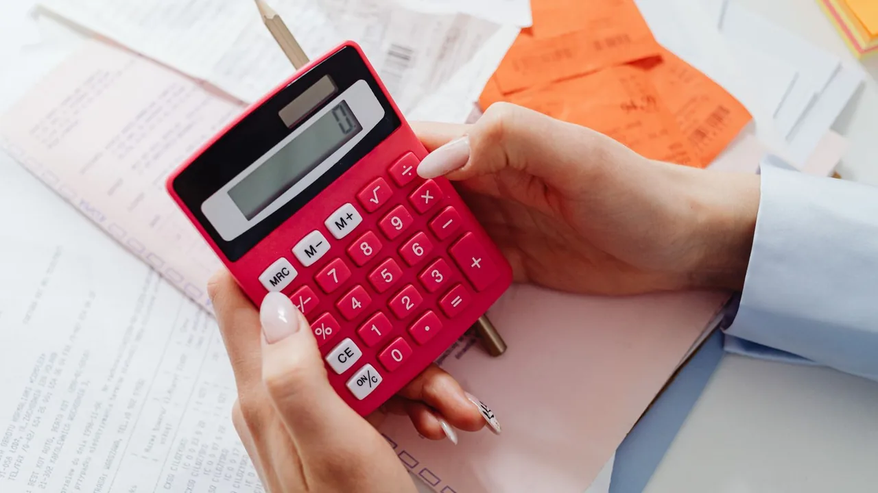 Des mains tiennent une calculatrice rose au-dessus de reçus et de documents posés sur un bureau