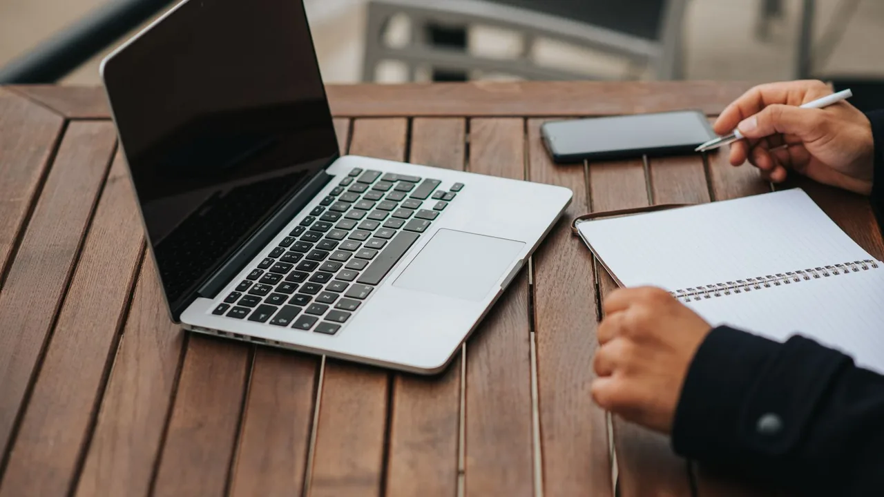 Ordinateur portable ouvert sur une table en bois, une main tenant un stylo près d'un carnet à spirale et d'un téléphone