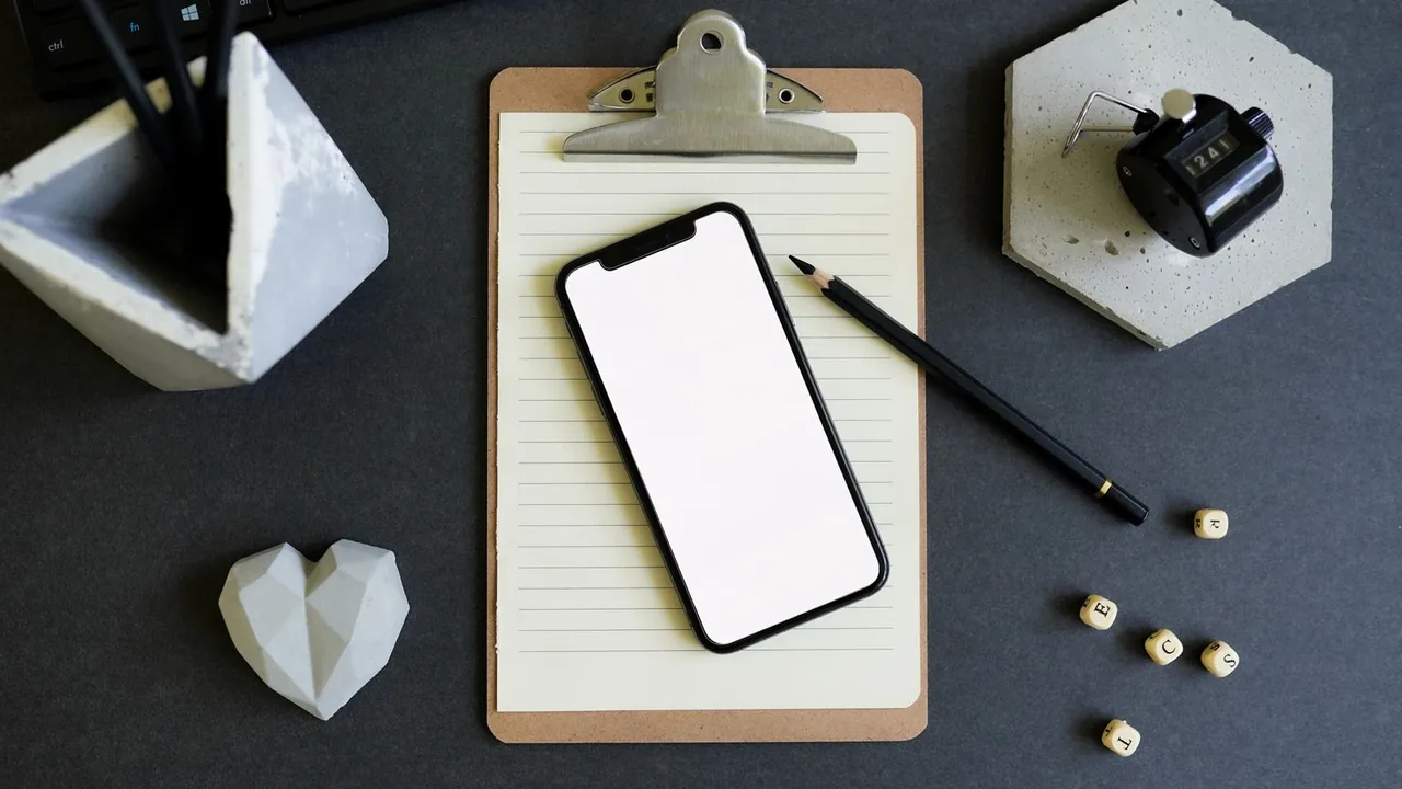 Smartphone posé sur un bloc-notes à pince, avec crayon, dés en bois et objets en béton sur un bureau gris foncé