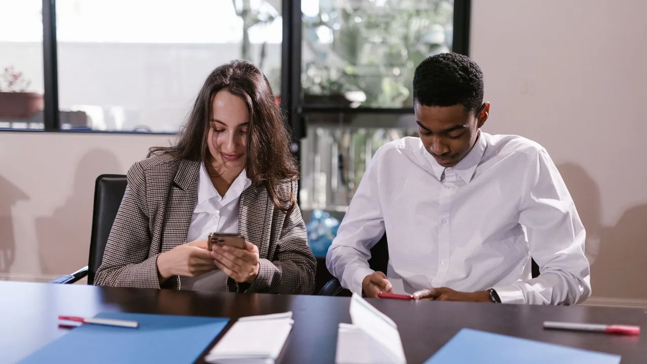 Deux jeunes professionnels assis à une table de réunion, concentrés sur leur smartphone, dossiers et feutres devant eux