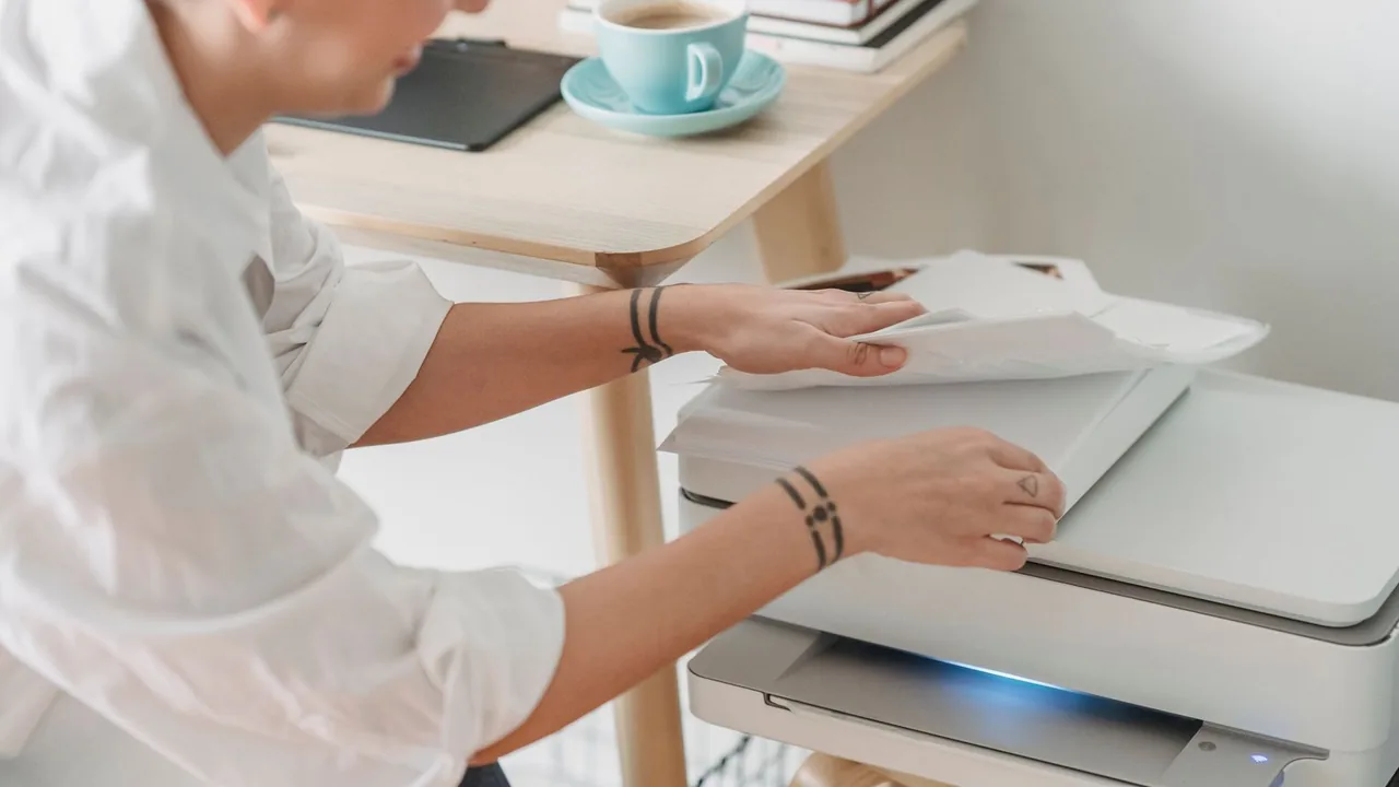 Une personne pose une feuille sur la vitre d'un scanner multifonction, une tasse bleue sur la table à côté.