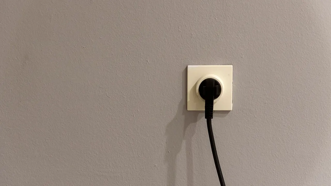 Une fiche noire branchée dans une prise murale encastrée sur un mur beige uni, son câble pendant vers le bas