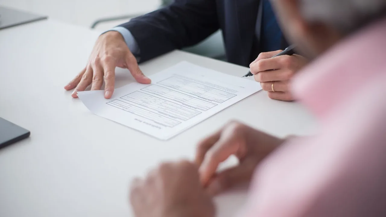 Un homme en costume pose la main sur un contrat et signe, face à un interlocuteur en chemise rose, flou au premier plan
