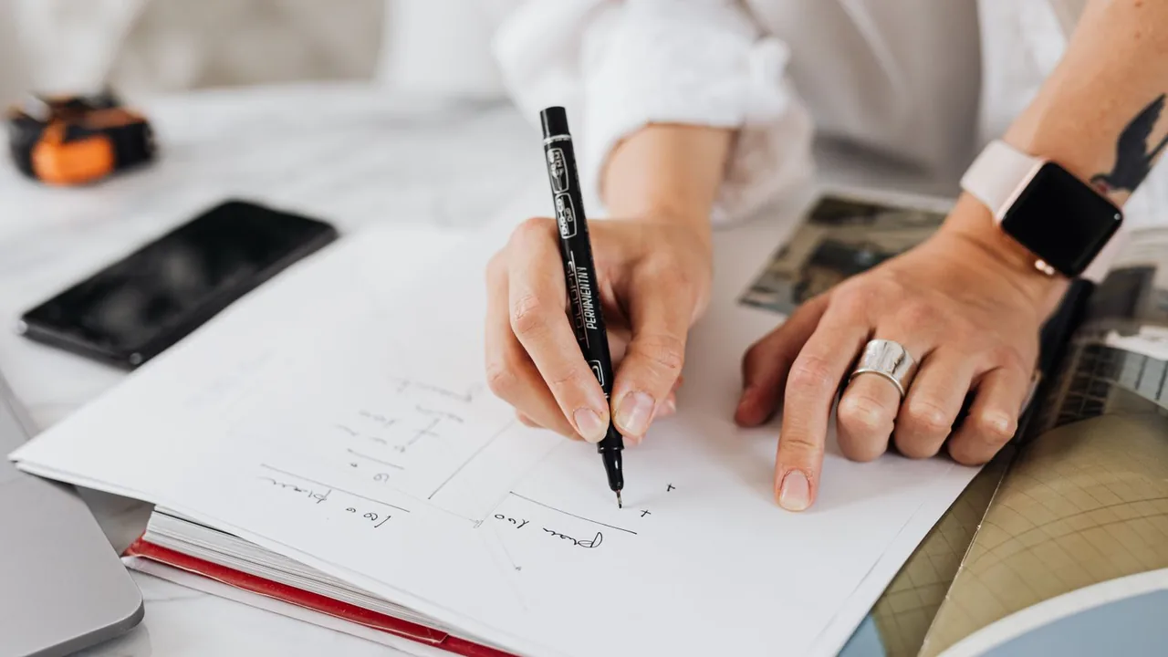 Gros plan d'une main qui dessine au stylo noir le schéma d'une page, près d'un ordinateur portable