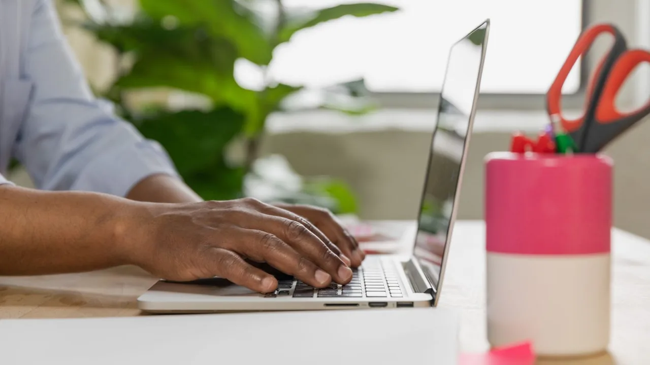 Mains tapant sur un ordinateur portable posé sur un bureau blanc, près d'une plante et d'un pot à crayons