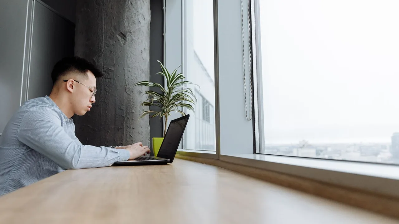 Homme concentré sur son ordinateur portable à une longue tablette en bois près d'une grande fenêtre