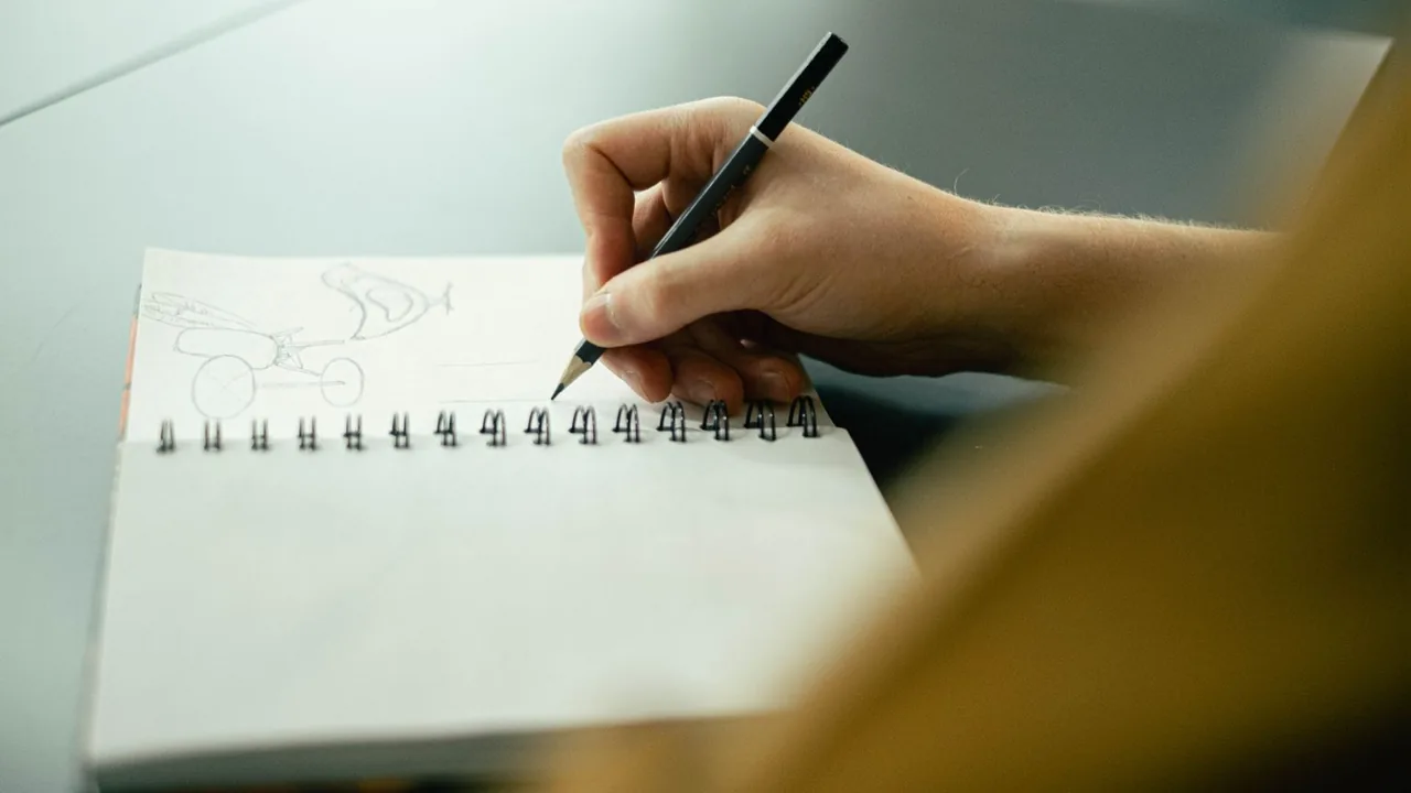 Main dessinant au crayon dans un carnet à spirale posé sur un bureau