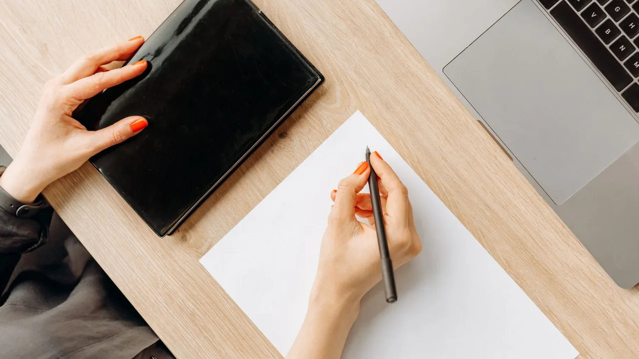 Une main tient un stylo au-dessus d'une feuille blanche, un ordinateur portable et un carnet noir sur un bureau clair