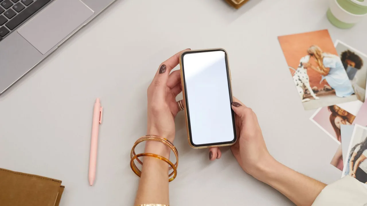 Une femme tient un smartphone à l'écran vide au-dessus d'un bureau avec un ordinateur portable et des photos