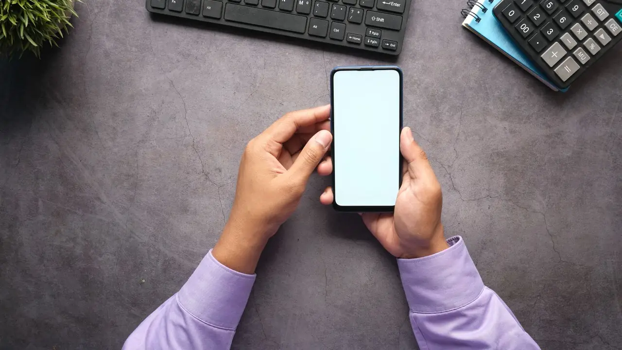 Vue de dessus des mains d'un homme tenant un smartphone, sur un bureau gris avec clavier, carnet et calculatrice