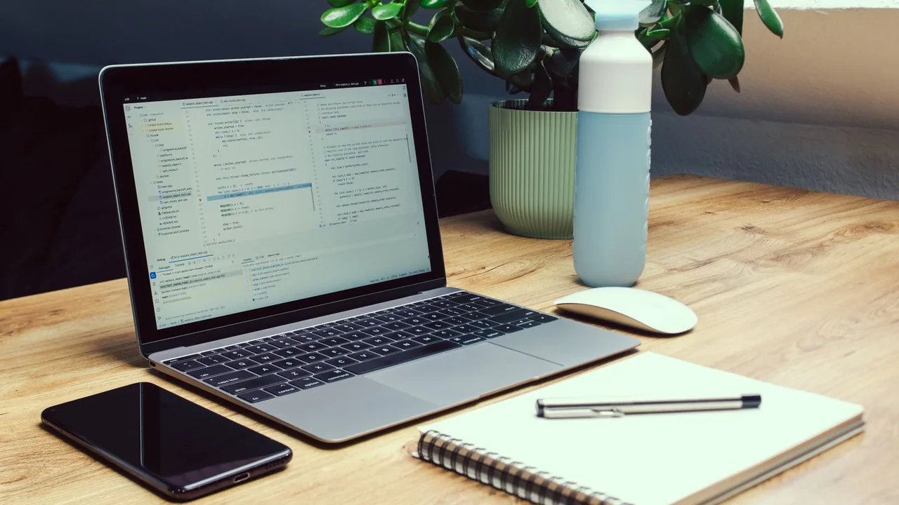Ordinateur portable ouvert à côté d'un smartphone et d'un carnet sur un bureau en bois, avec une plante et une gourde