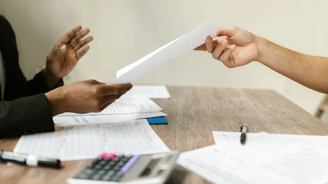 Des mains se passent des documents sur une table, avec une calculatrice et des feuilles de chiffres