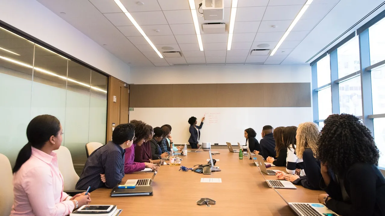 Une collaboratrice présente au tableau au fond d'une longue table de réunion, des ordinateurs portables devant les participants
