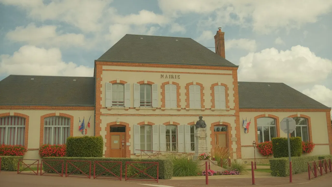 Une mairie de village aux murs crème et aux encadrements de briques rouges, entourée de haies et de massifs de fleurs