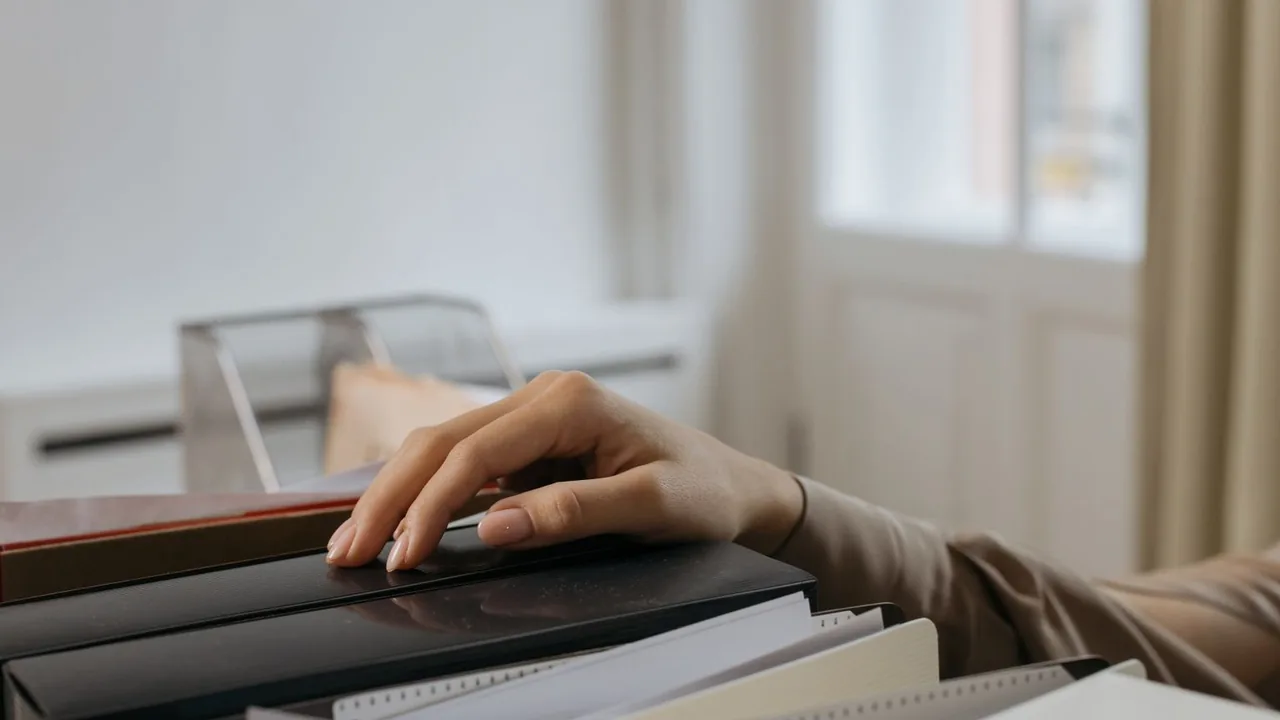 Une main parcourt une pile de classeurs et de dossiers posés sur un bureau