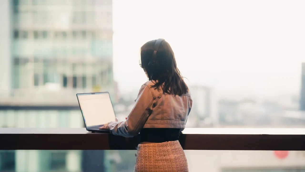 Femme debout devant son ordinateur portable posé sur un garde-corps, face à une vue urbaine brumeuse
