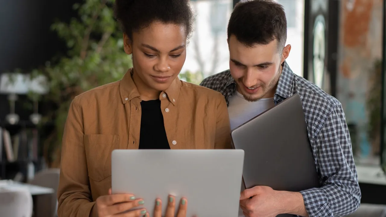 Deux collègues regardent ensemble l'écran d'un ordinateur portable, l'un tenant un second portable sous le bras