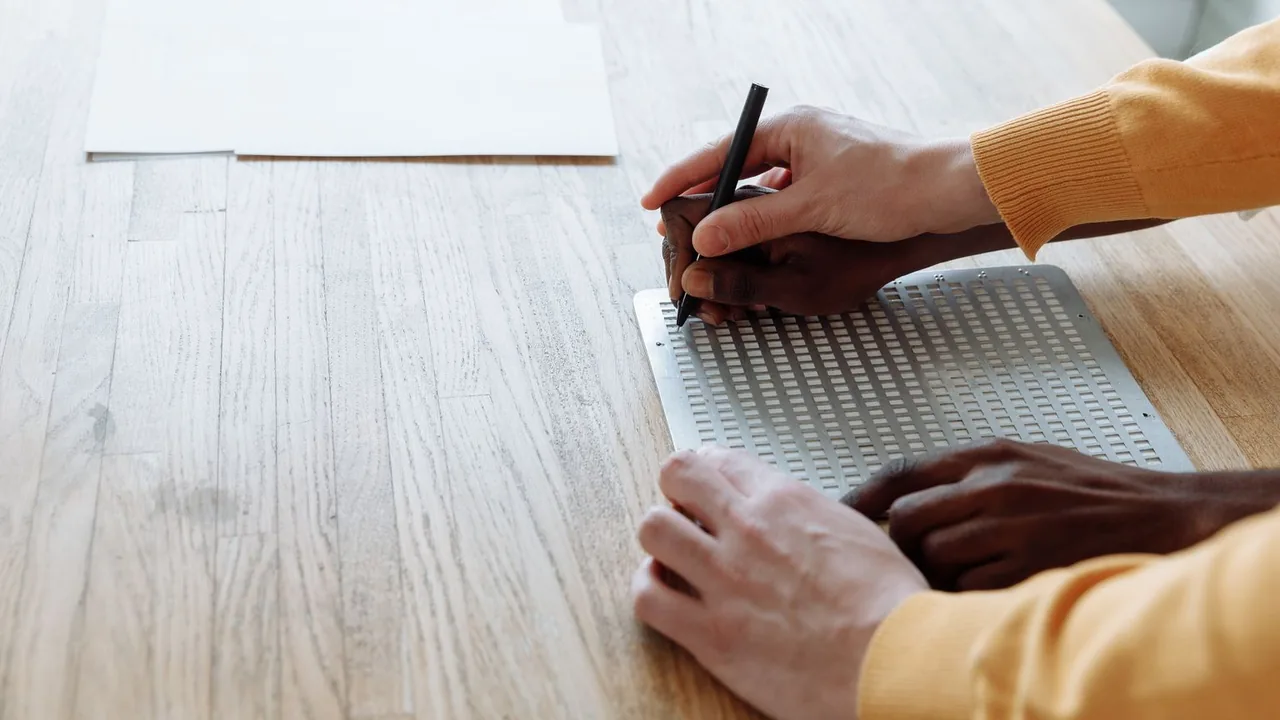 Des mains écrivent en braille avec un poinçon sur une planchette posée sur une table en bois