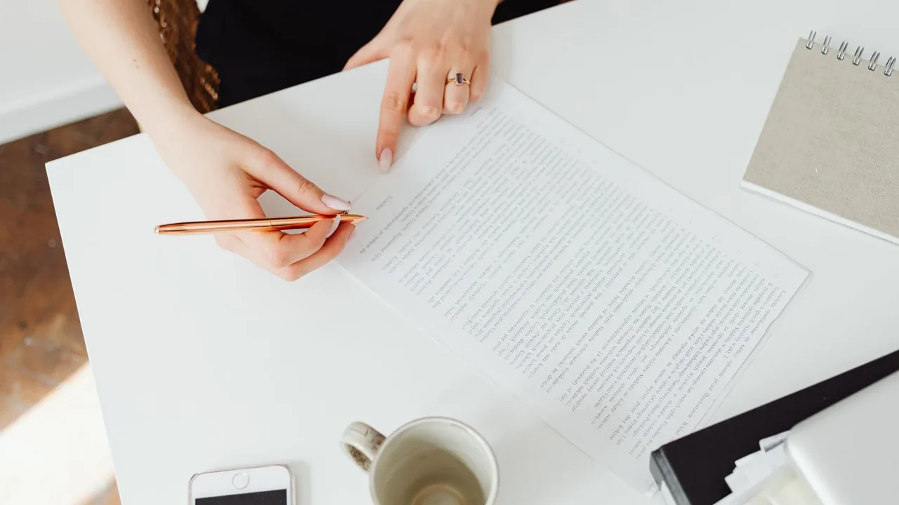 Deux mains, l'une tenant un stylo cuivré, l'autre pointant un contrat imprimé posé sur un bureau blanc