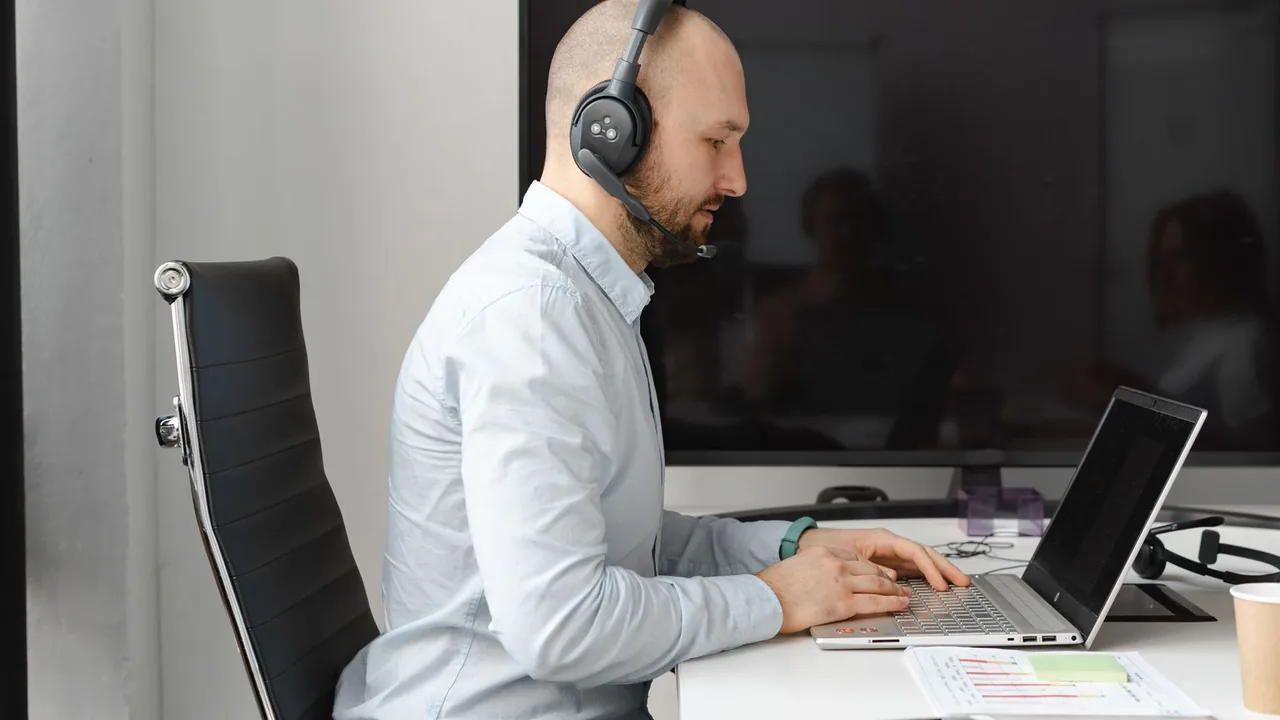 Un conseiller en chemise claire, casque-micro sur la tête, tape sur son ordinateur portable à un bureau blanc