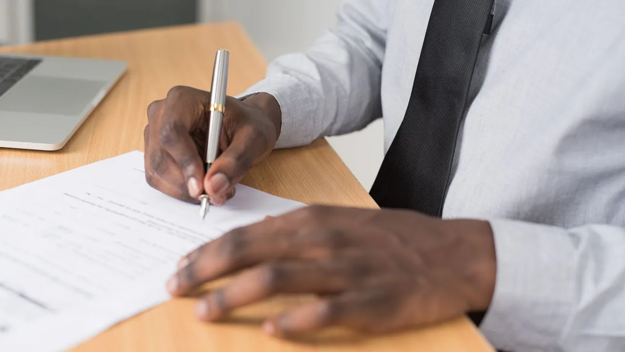 Un homme en chemise grise signe un document avec un stylo plume sur un bureau en bois, un ordinateur portable à côté