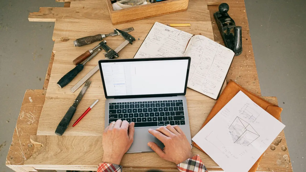Vue de dessus des mains d'un menuisier sur un ordinateur portable posé sur son établi, entouré d'outils et d'un croquis