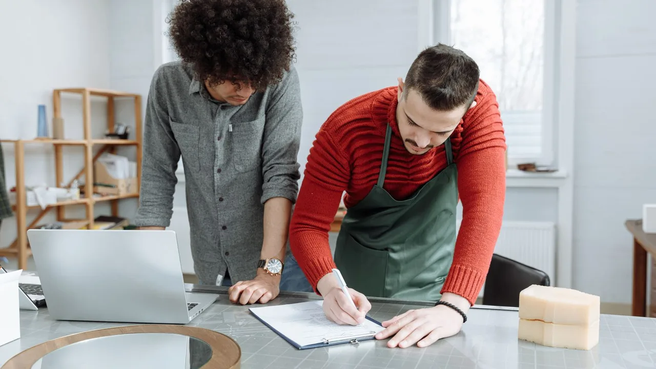 Deux artisans debout à leur plan de travail : l'un remplit un formulaire sur un presse-papiers près d'un ordinateur portable