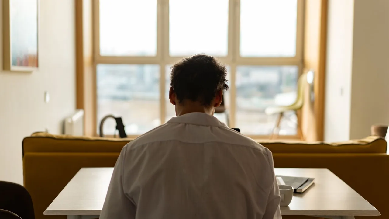 Homme de dos assis à une table devant une grande fenêtre dans un salon lumineux