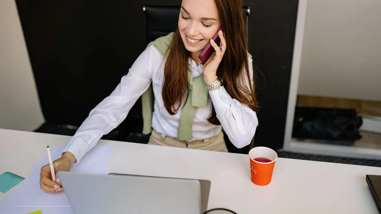Une femme souriante au téléphone prend des notes sur une feuille devant son ordinateur portable, un gobelet orange à côté.