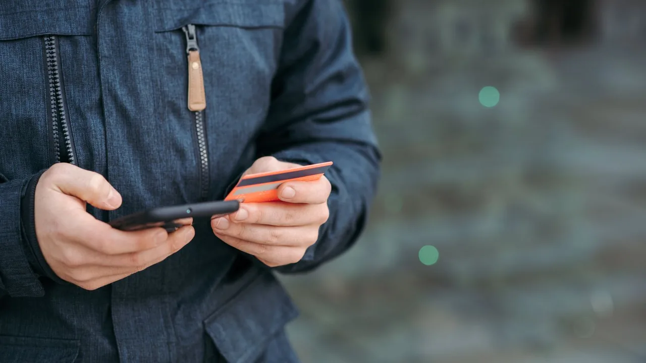 Des mains tiennent un smartphone et une carte bancaire pour un paiement, en extérieur