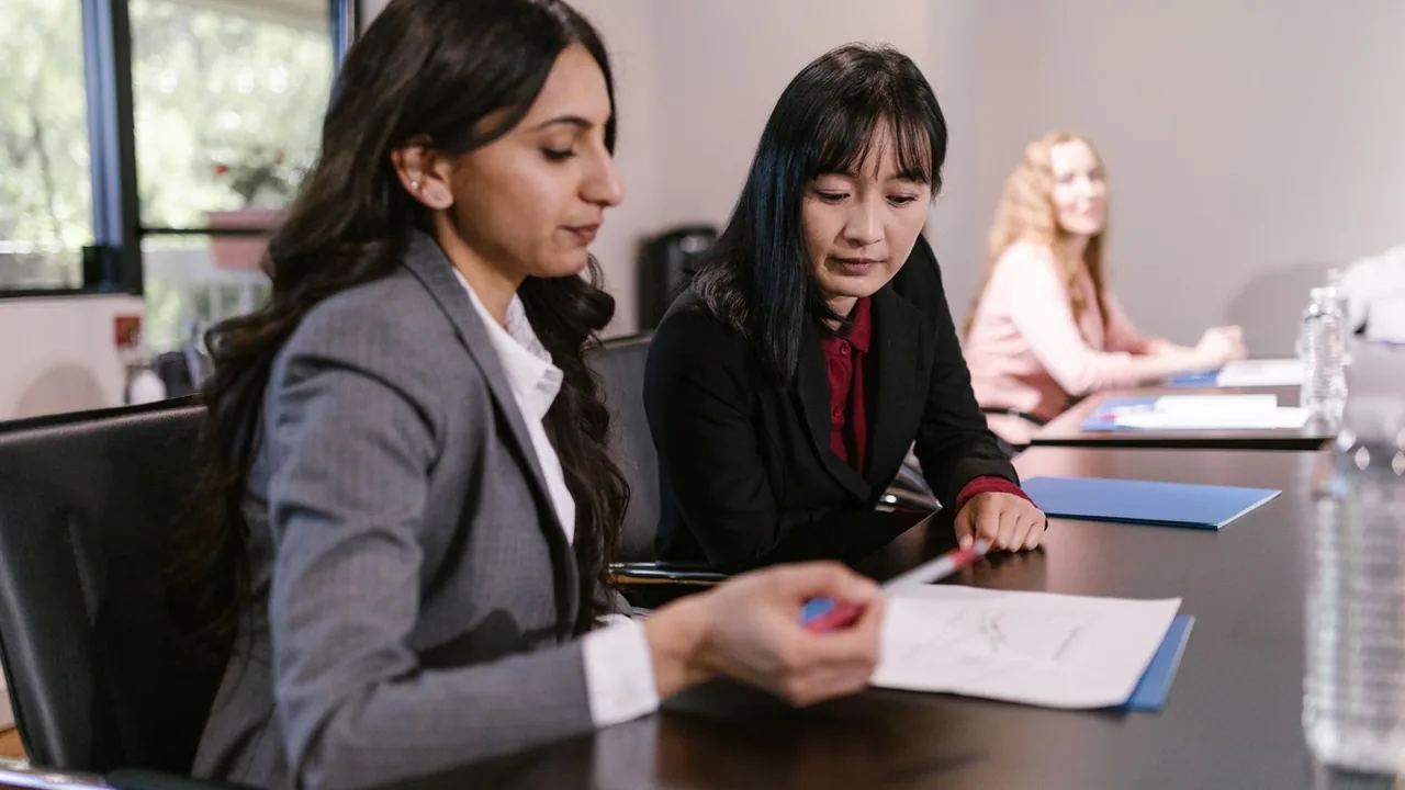 Deux femmes en blazer examinent un document autour d'une table de réunion, une troisième au fond de la salle