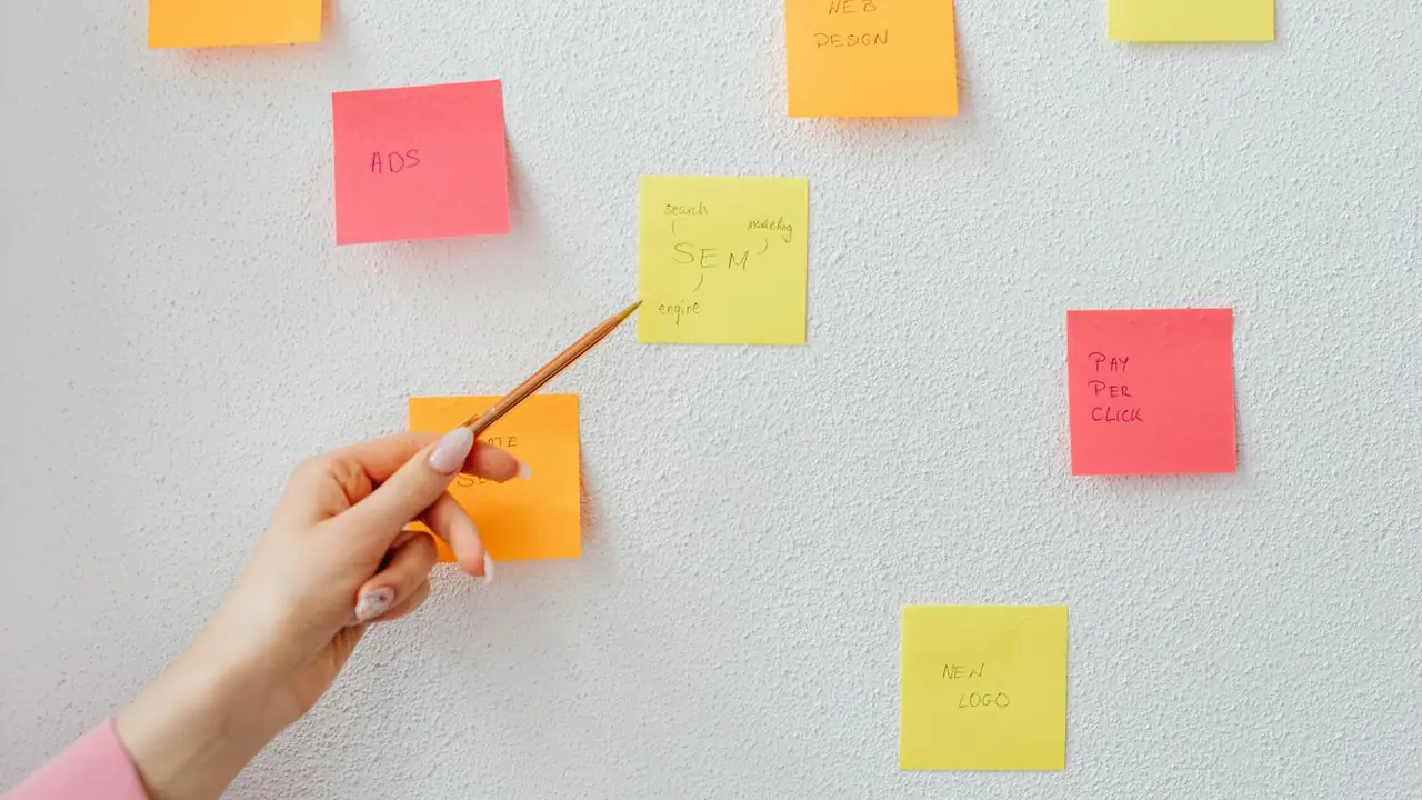Une main désigne des notes autocollantes colorées fixées sur un mur blanc pour planifier des contenus