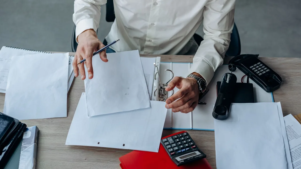 Vue de dessus d'un professionnel en chemise blanche qui trie des documents, calculatrice et téléphone sur le bureau