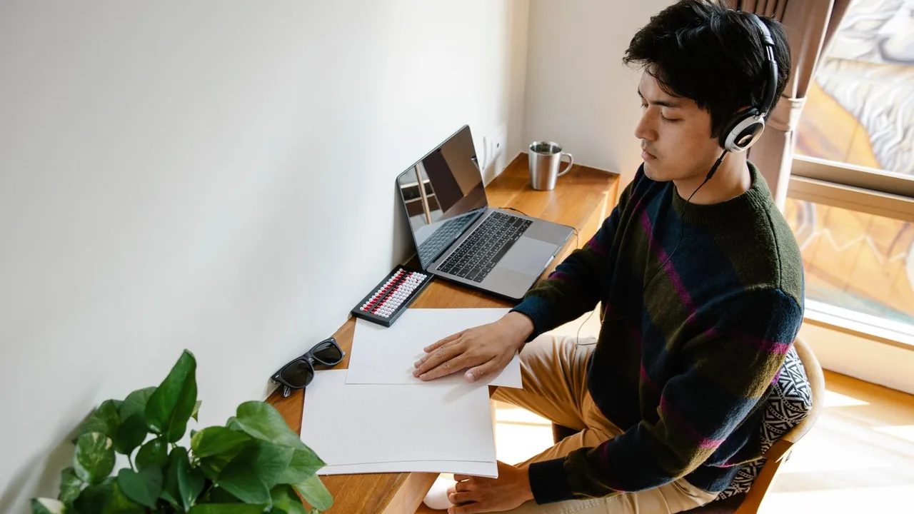 Un jeune homme avec un casque audio travaille à un bureau en bois, des feuilles et un ordinateur portable devant lui
