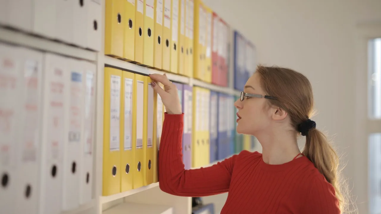 Une femme cherche un dossier parmi des classeurs colorés alignés sur une étagère de bureau.