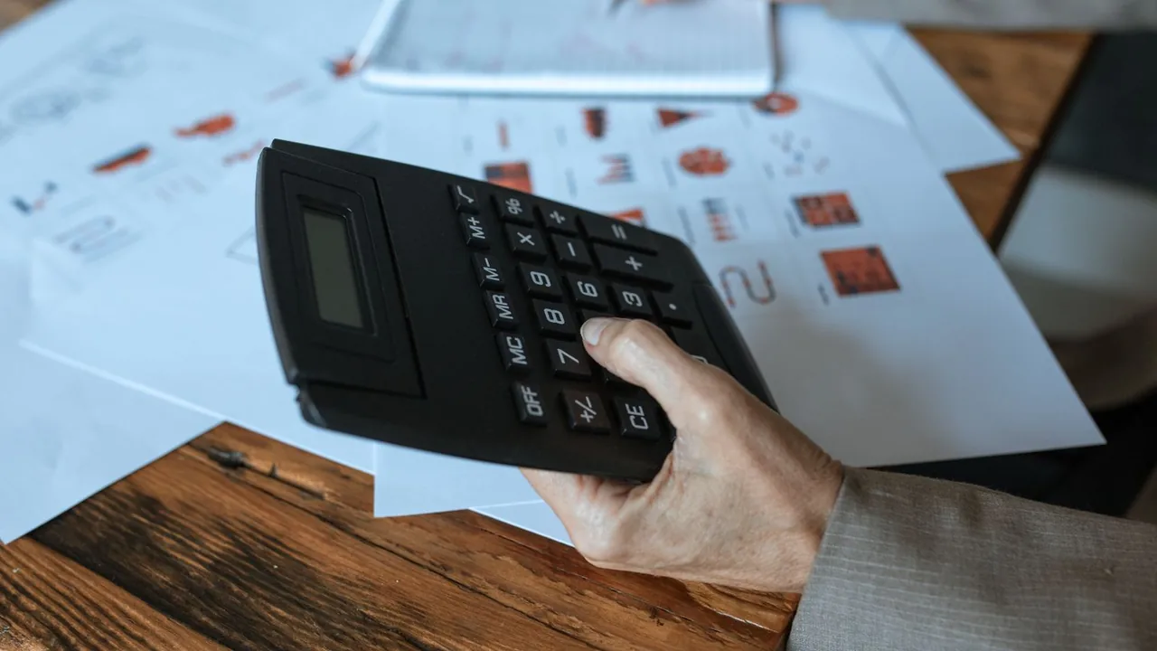 Une main utilise une calculatrice sur un bureau en bois couvert de documents