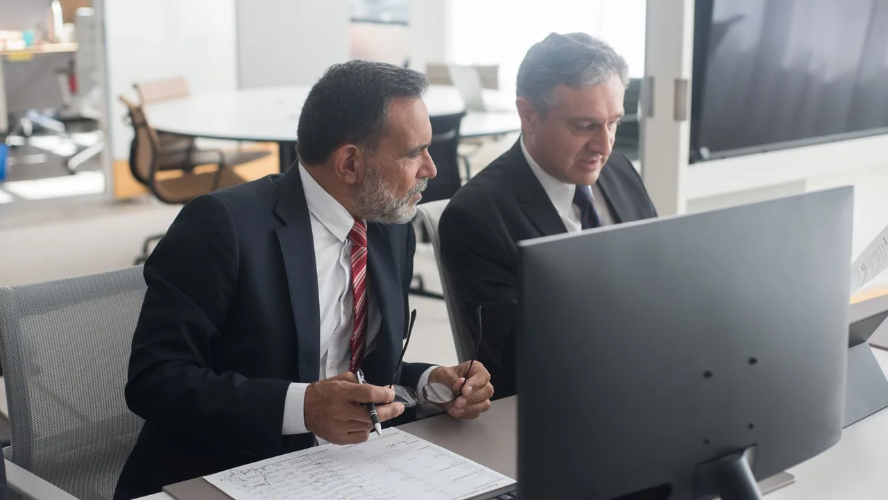Deux hommes en costume consultent un dossier et un écran à leur poste de travail