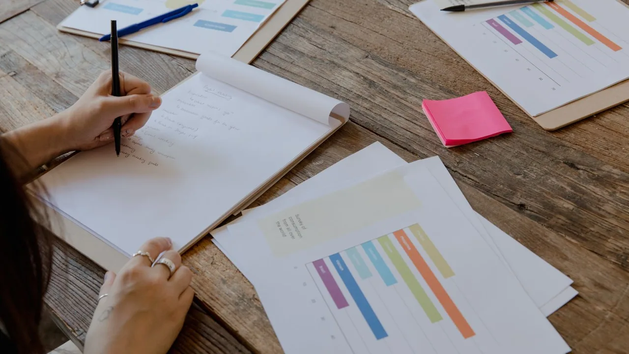 Mains qui écrivent sur un carnet, à côté de rapports imprimés aux graphiques colorés sur une table en bois