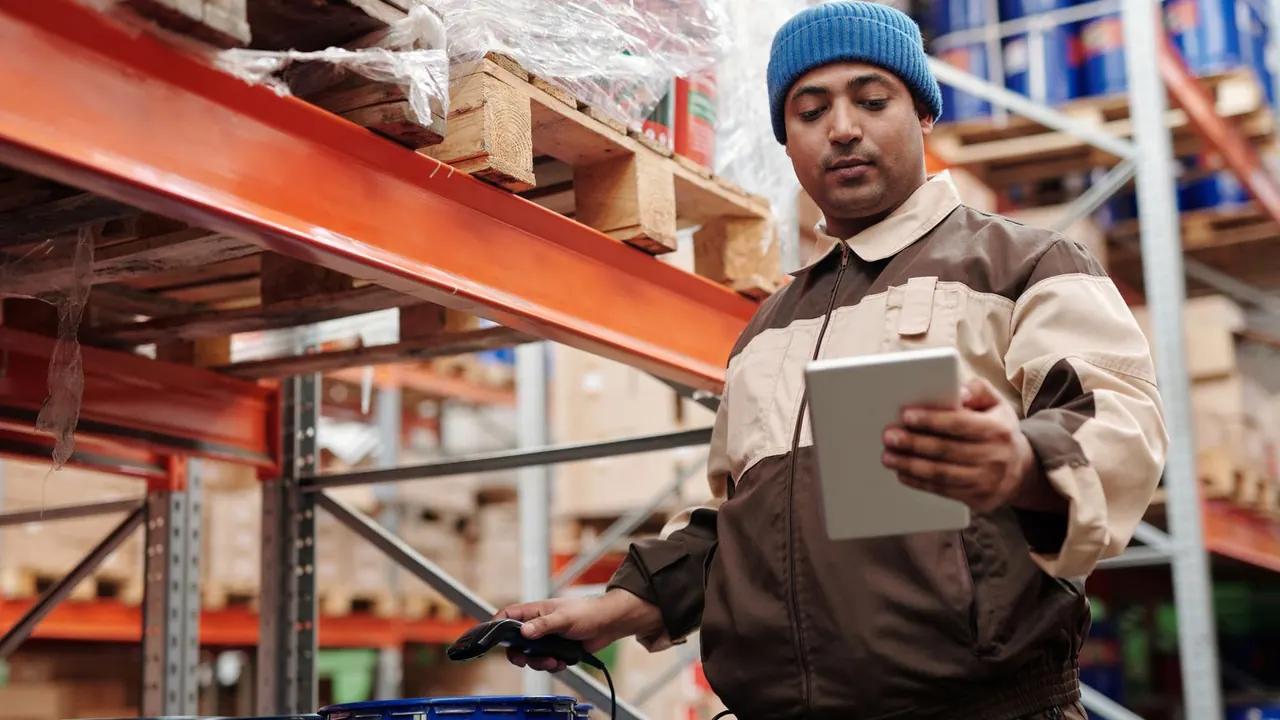 Un employé d'entrepôt consulte une tablette entre de hauts rayonnages.