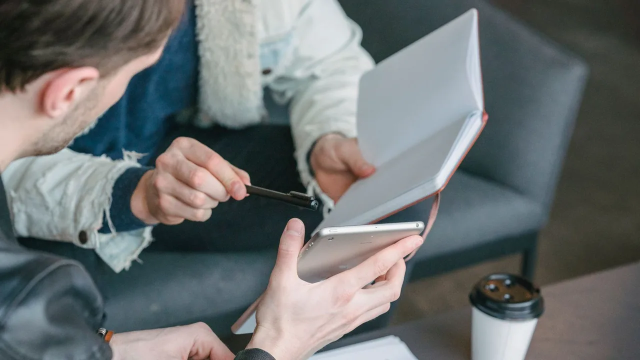 Deux collègues discutent autour d'un carnet et d'une tablette, un gobelet de café posé à côté.