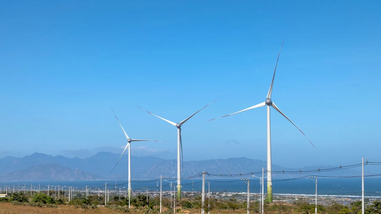 Rangée d'éoliennes devant des montagnes et des lignes à haute tension.