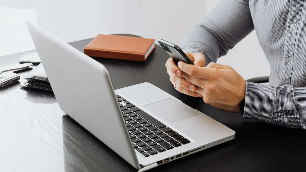 Un homme tient son smartphone à son bureau, devant un ordinateur portable ouvert et un carnet
