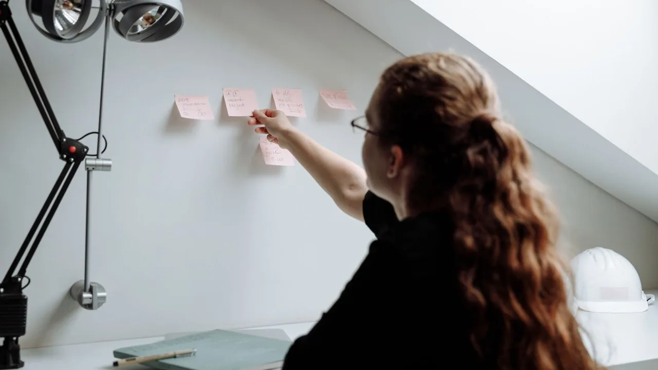 Une femme, de dos, colle des post-it roses sur un mur blanc, un casque de chantier posé sur le bureau