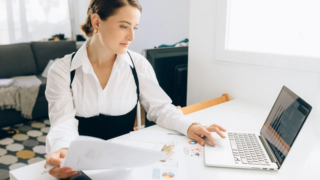 Une femme à son bureau parcourt un rapport imprimé en consultant son ordinateur portable