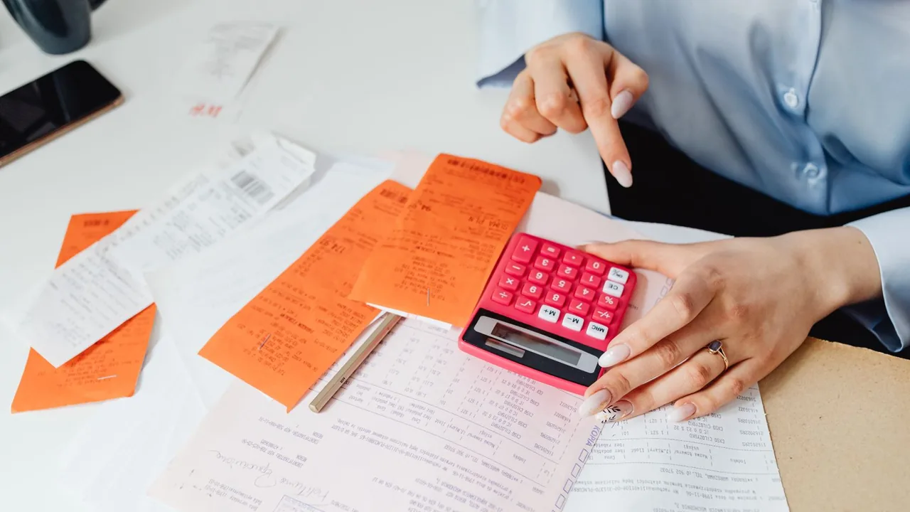 Une main pointe un reçu posé sur des factures étalées à côté d'une calculatrice rose, lors d'un contrôle
