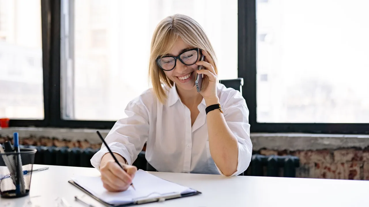 Femme au téléphone qui prend des notes sur un bloc à son bureau, pendant l'appel de relance d'un client