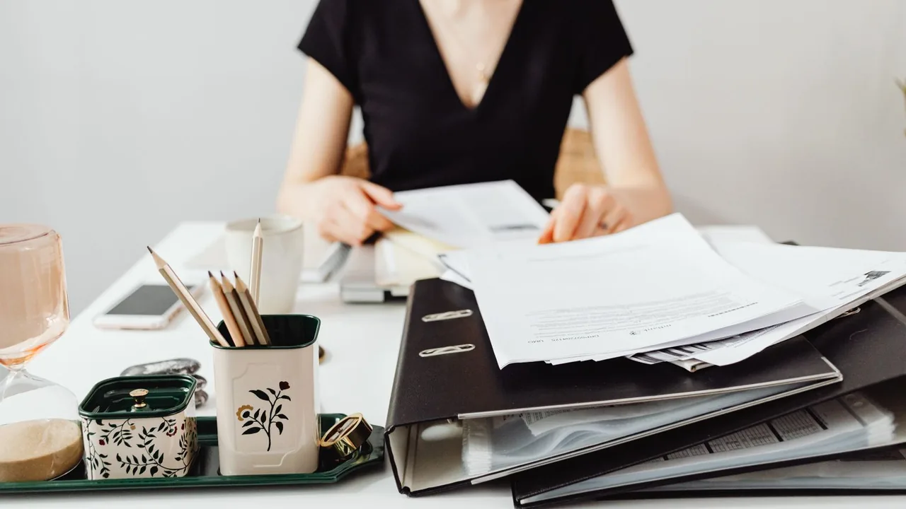 Femme au bureau devant une pile de classeurs et de feuilles à ranger, pot à crayons à côté