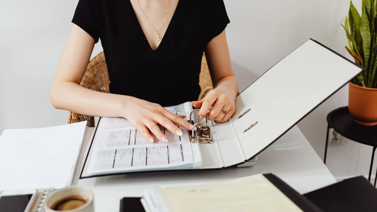 Une femme feuillette un classeur de factures ouvert sur un bureau clair, une tasse de café à côté
