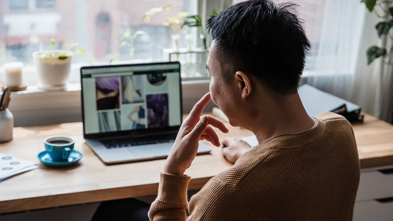 Un homme en pull relit le contenu affiché sur son ordinateur portable, une tasse de café posée sur le bureau