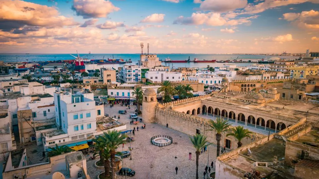 Vue sur la médina de Sousse : place, remparts et arcades de la Grande Mosquée, avec le port à l'horizon au coucher du soleil